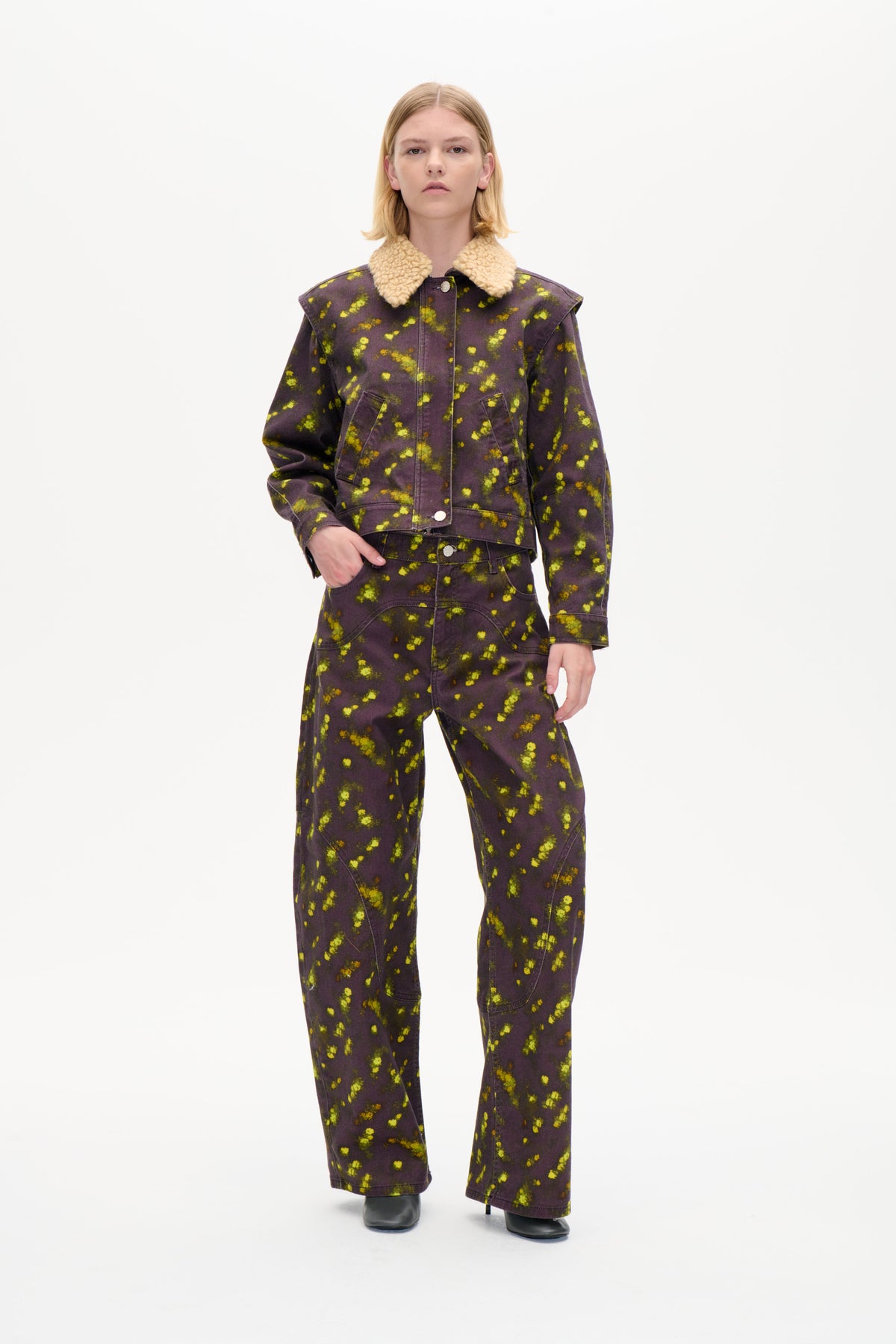 Model trägt eine dunkelpflaumenfarbene Jacke mit cremefarbenem, strukturiertem Kragen und einer passenden, weit geschnittenen Hose, beide sind mit leuchtend gelb-grünen Markierungen gemustert.