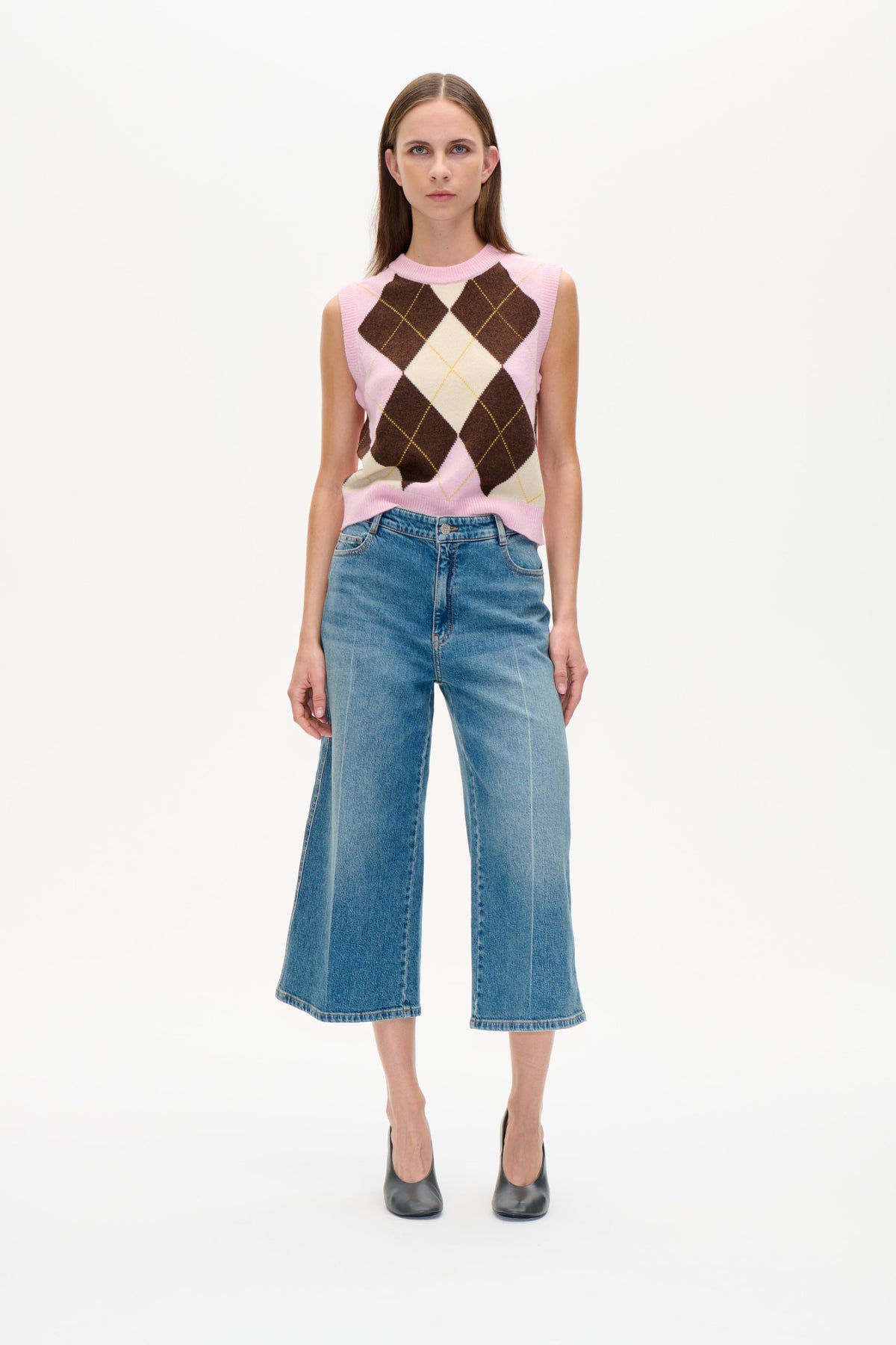 Model wearing a pink sleeveless knit vest with brown and cream argyle diamonds, paired with blue wide-leg jeans.
