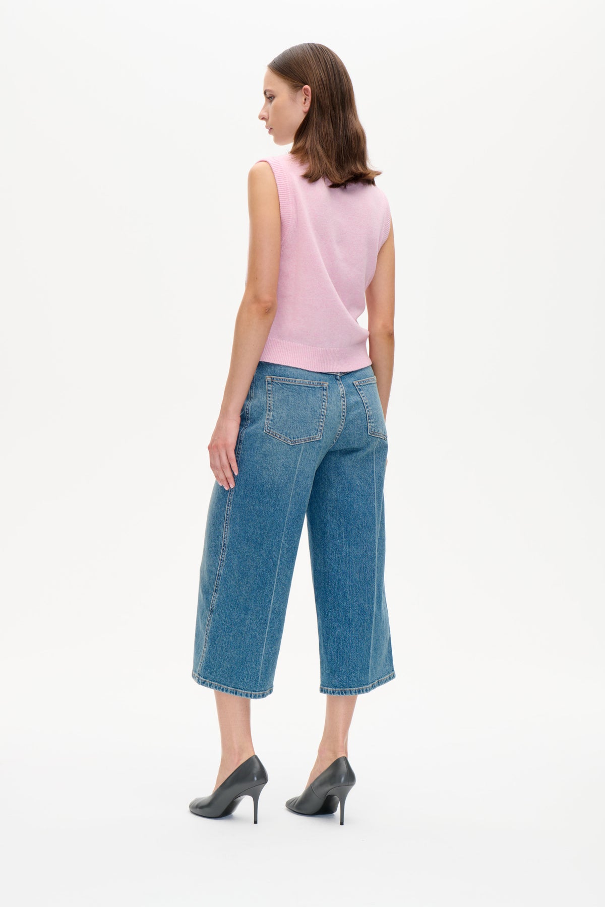 Back three-quarter view of a model wearing a pale pink sleeveless knit vest with ribbed trim and blue wide-leg cropped jeans.