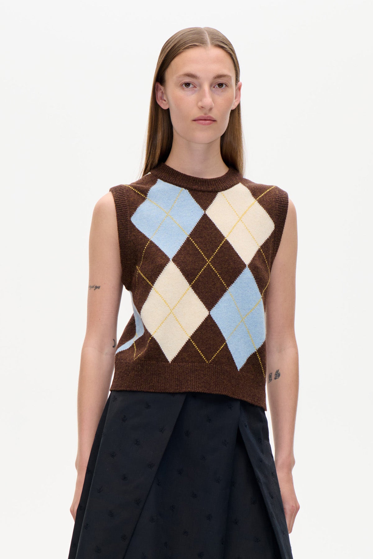 Vue de face d'un gilet sans manches en maille marron, à grands losanges argyle bleu pâle et crème, porté avec une jupe noire.