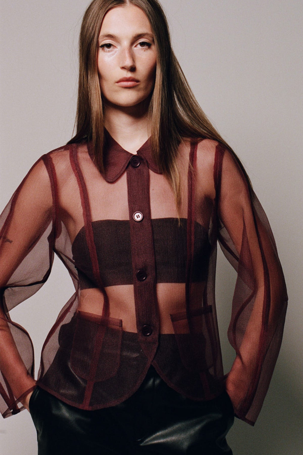 Campaign portrait of a model wearing a sheer burgundy and brown checked button-up shirt over a black top