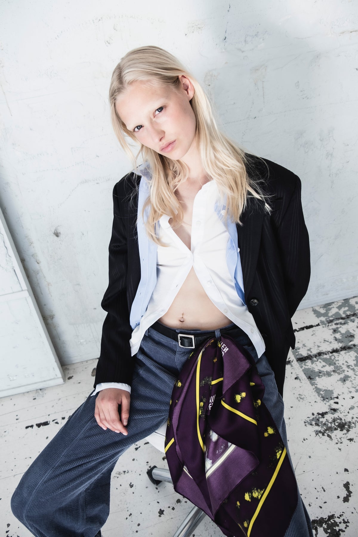 Kampagnenportrait eines Models in schwarzem Blazer, weißem Hemd mit Kragen und hellblauer, feingestreifter Weste; dazu ein violetter Blumenschal zu dunkelblauen Hosen.