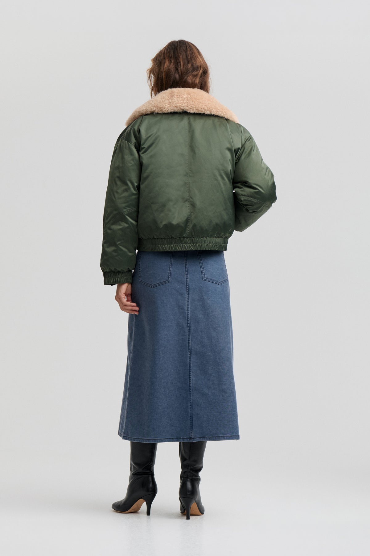 Back view of a model wearing an olive green jacket with a wide beige shearling-style collar, paired with a blue denim midi skirt.
