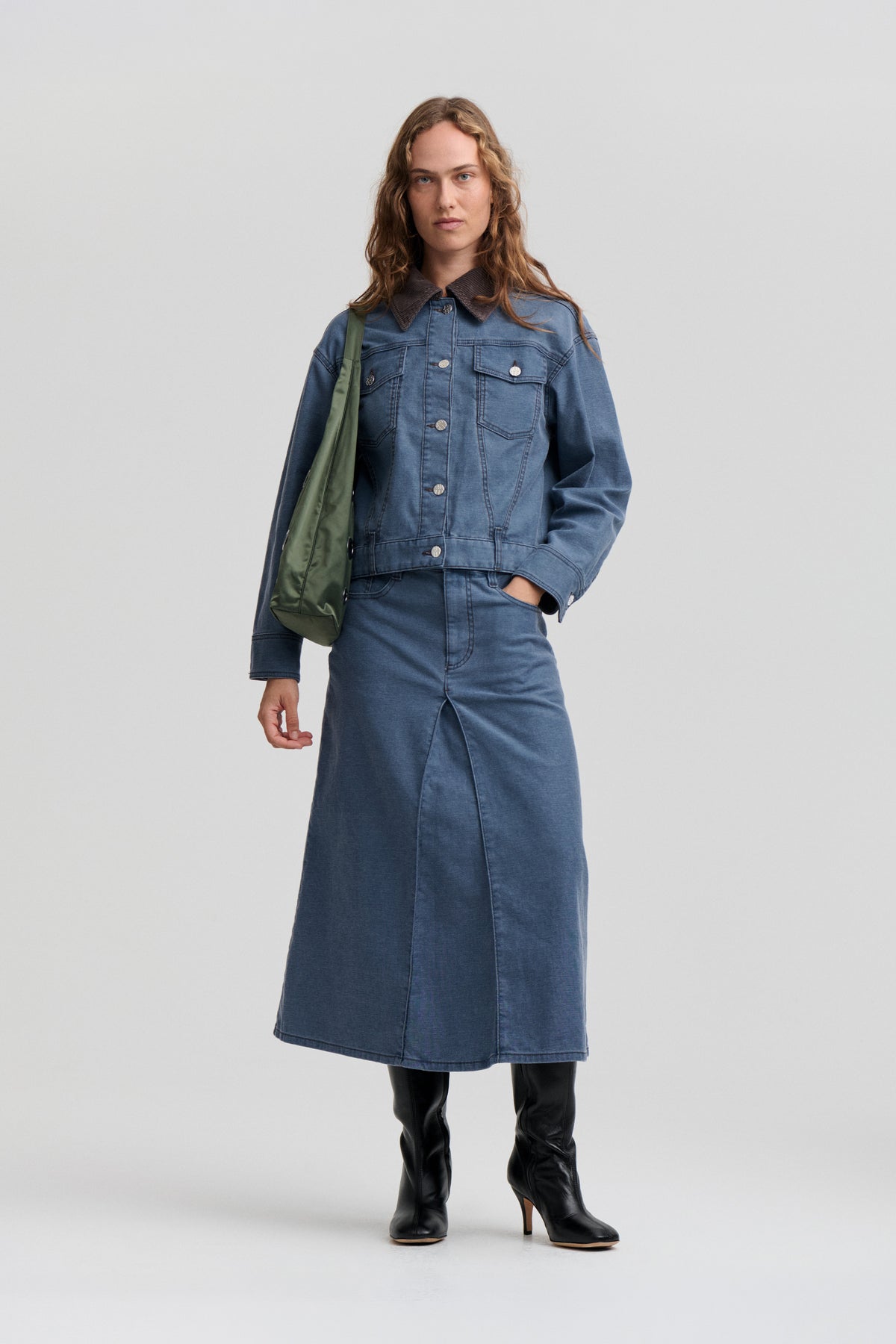 Model wearing a blue denim jacket with a dark corduroy collar over a matching long denim skirt, carrying a green shoulder bag.