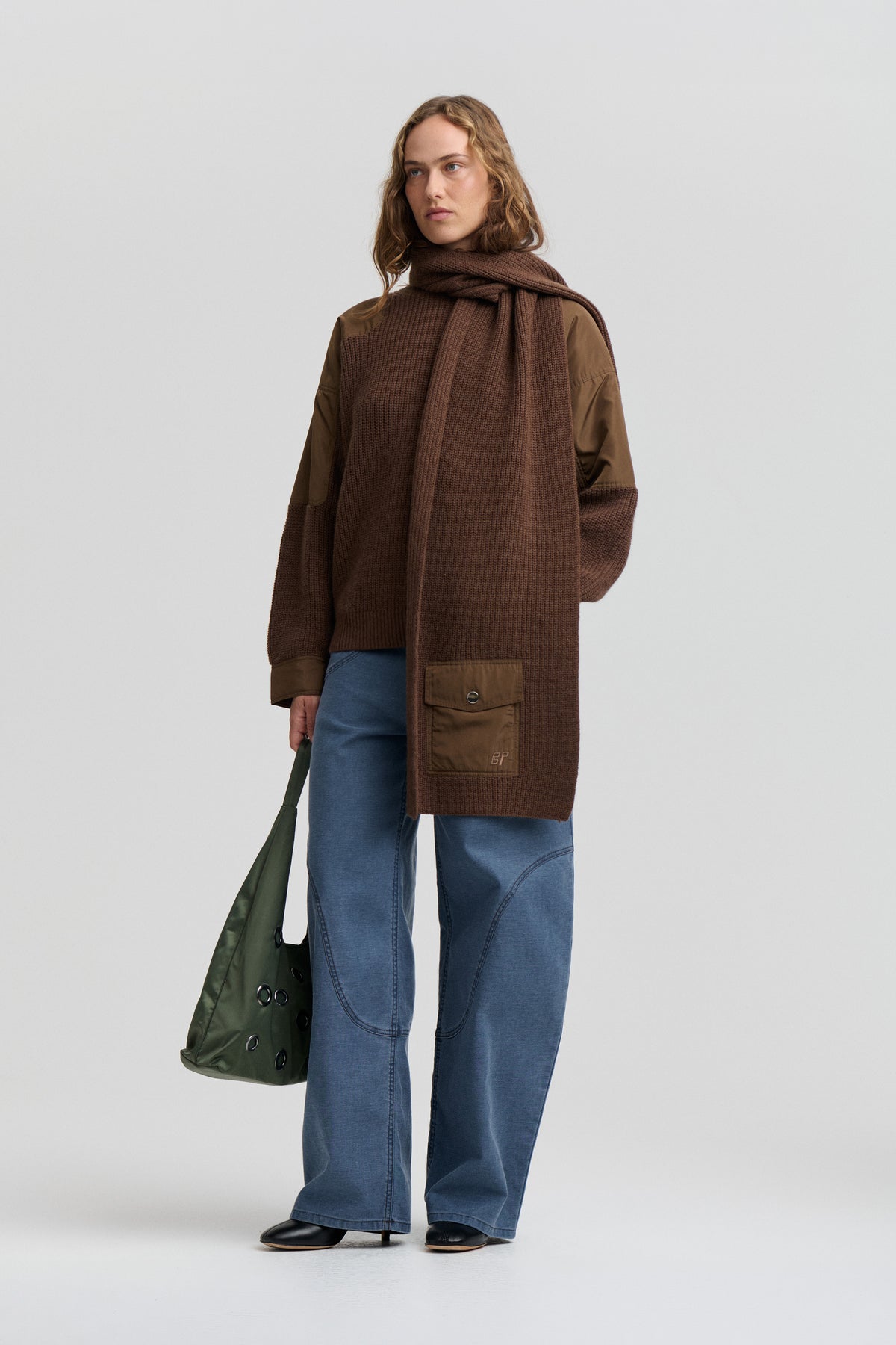Model wearing a long brown textured knit coat with a matching draped scarf, contrast fabric panels, blue wide-leg jeans and a dark green bag.