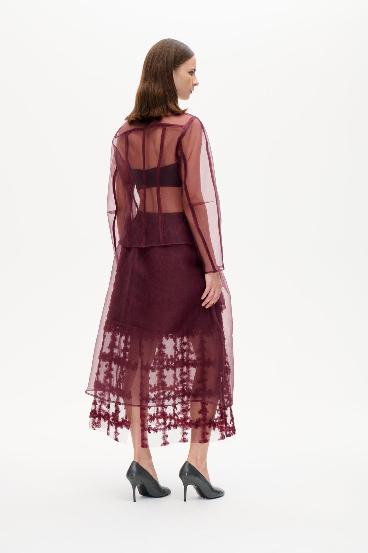 Model in a sheer plum long-sleeved blouse with dark panel seams, layered over a dark outfit and a sheer skirt with burgundy floral detailing.