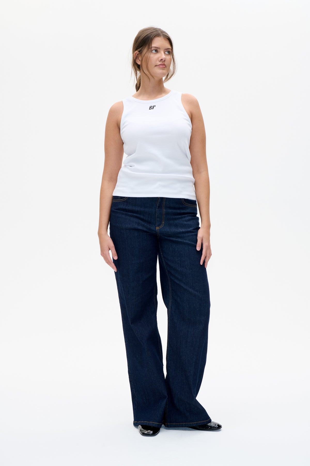 Model wearing a white sleeveless ribbed top with a small dark chest logo and dark blue wide-leg jeans, shown from the front.