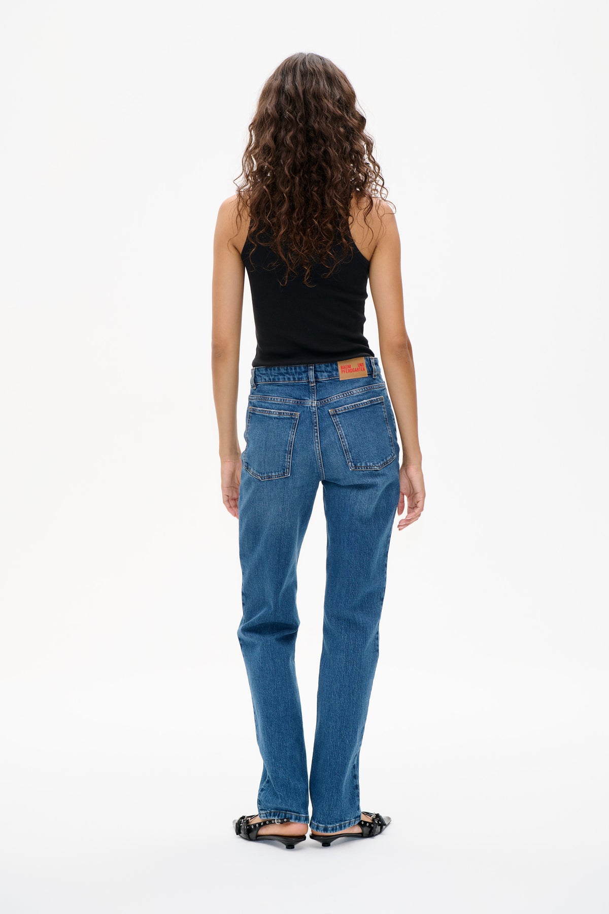 Achteraanzicht van een model dat een zwarte mouwloze top, blauwe rechte jeans met opgezette achterzakken en zwarte schoenen met bandjes draagt.