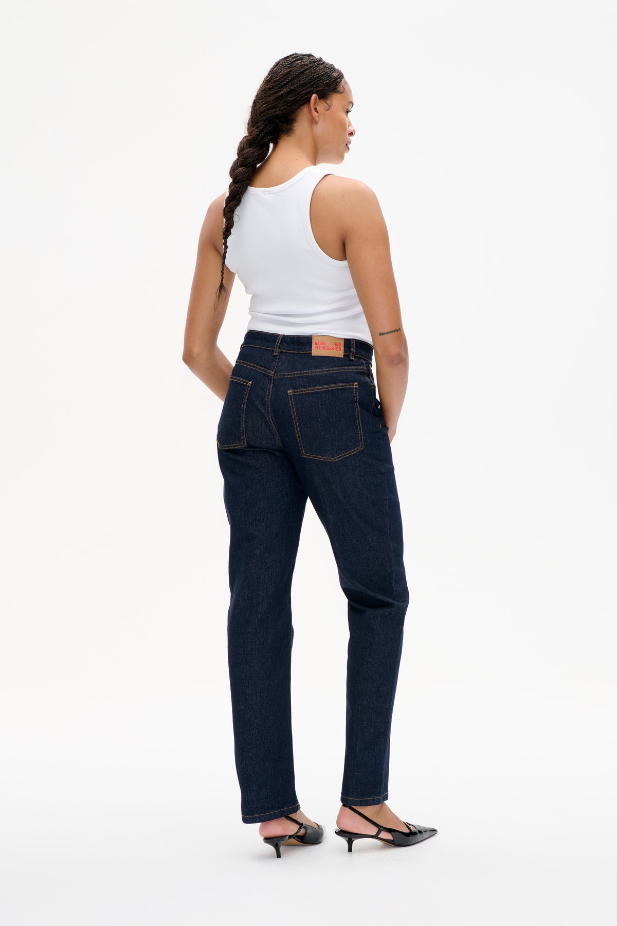 Model van achteren gezien draagt een witte mouwloze top en een rechte jeans in donker indigoblauw met contrasterende stiksels en achterzakken.