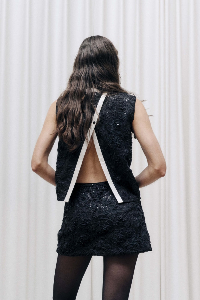 Back view of a model wearing a black textured sleeveless top with an open back and white crossed straps, paired with a matching mini skirt.