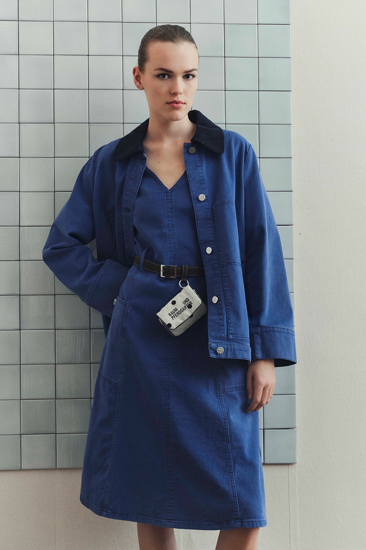 Model wearing a blue button-front jacket with a dark collar over a matching V-neck dress, styled with a black belt and crossbody pouch.