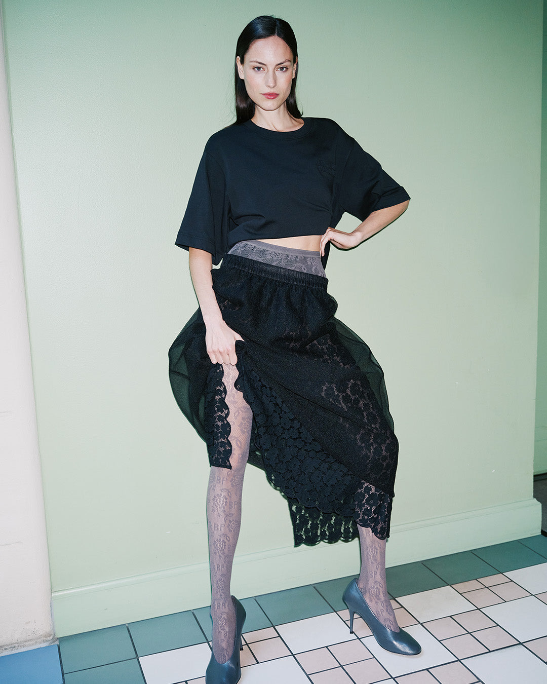 Model wearing a black cropped T-shirt and sheer floral lace skirt with patterned tights and blue heels.
